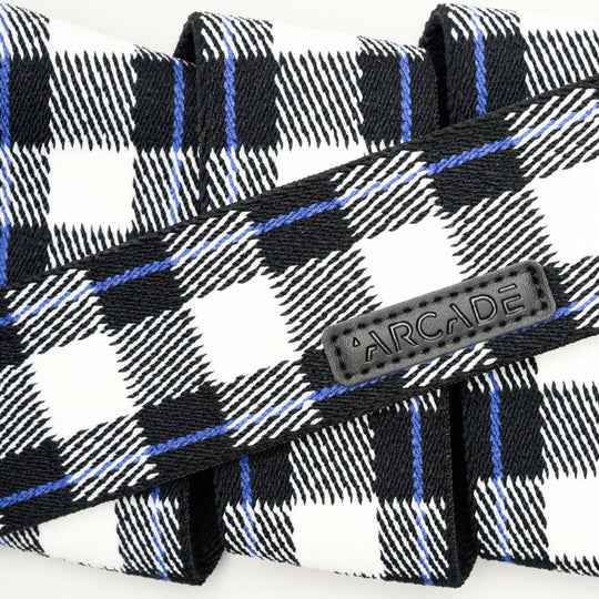 A close-up of the Plaid belt by Arcade Belts, featuring black, white, and blue plaid with a black ARCADE patch label. The elastic stretch belt is folded to show its woven texture and comfort—perfect for travel.